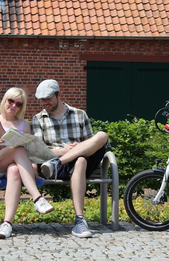 Radfahrer Bücken Drei Menschen sitzen auf einer Bank in sommerlicher Kleidung. Zwei Fahrräder stehen daneben. Eine Person hält eine Karte in den Händen. Im Hintergrund ist eine Backsteinmauer mit grünem Tor zu sehen.