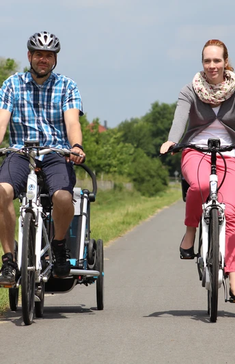 Zwei Radfahrer fahren auf einem asphaltierten ländlichen Weg in einer grünen, bewaldeten Landschaft.