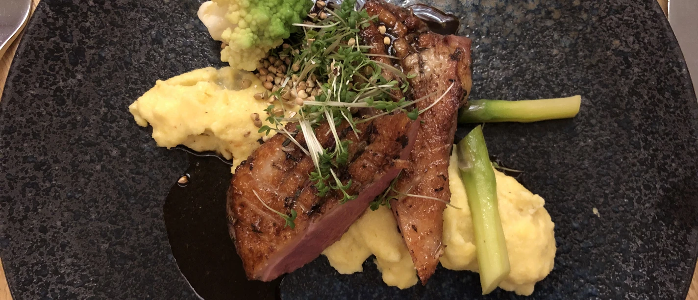 Duck breast on mashed potatoes with green asparagus spears and romanesco on a blue plate.