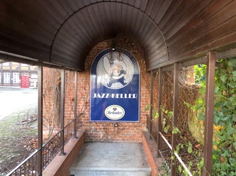 Eingang zum Jazz Keller Nienburg mit einem gewölbten Holzdach, umgeben von Backstein und grünen Pflanzen.