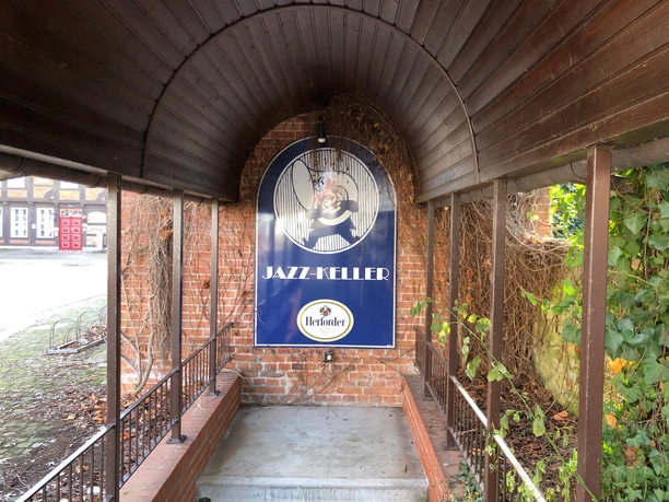 Jazz Keller Nienburg Eingang zum Jazz Keller Nienburg mit einem gewölbten Holzdach, umgeben von Backstein und grünen Pflanzen.