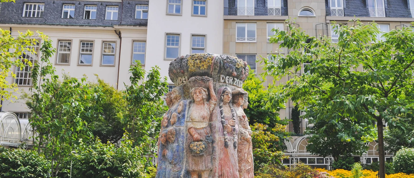 Der Frauenbrunnen zeigt zehn lebensgroße Frauenfiguren "die Frauen Kölns im Wandel der Zeiten“, umgeben von üppigem Grün, vor urbaner Kulisse.