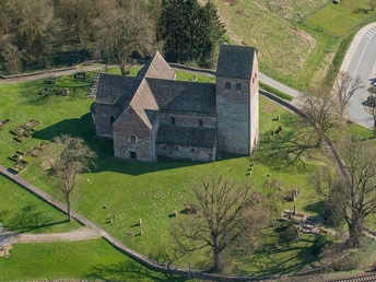 Luftaufnahme einer alten Kirche mit rechteckigem Turm, umgeben von grüner Wiese und Bäumen.