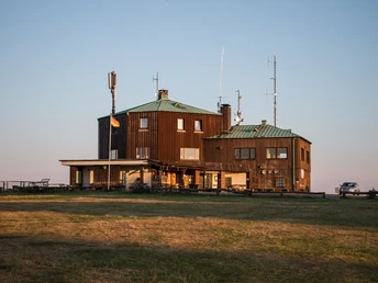 Großes, teils hölzernes Gebäude mit grünen Dächern, Antennen auf dem Dach, auf freiem Feld.
