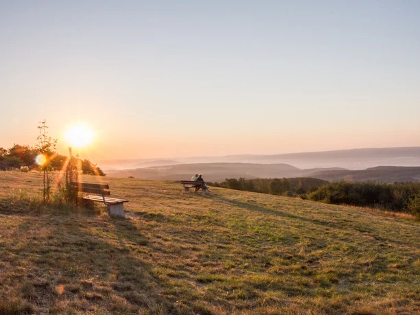 Sonnenaufgang auf dem Köterberg Sonnenaufgang über Hügeln am Köterberg; zwei Personen auf Bank, sanftes Morgenlicht, weite Fernsicht.