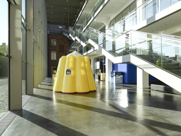 Gelbe Puddingform-Skulptur in modernem Glasgebäude mit reflektierendem Boden und Treppen.