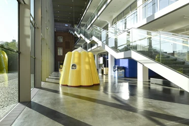 Das Puddingwunder in der Dr. Oetker Welt Gelbe Puddingform-Skulptur in modernem Glasgebäude mit reflektierendem Boden und Treppen.