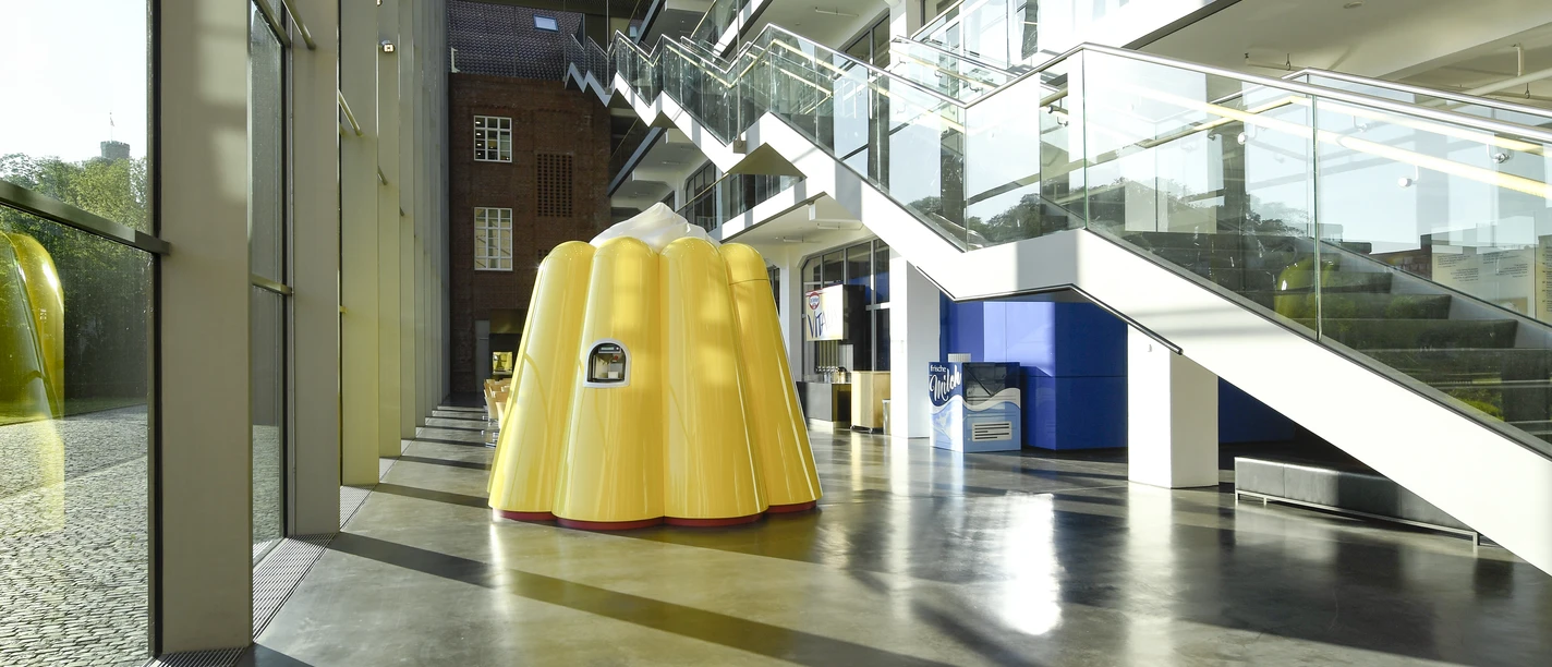 Das Puddingwunder in der Dr. Oetker Welt Gelbe Puddingform-Skulptur in modernem Glasgebäude mit reflektierendem Boden und Treppen.