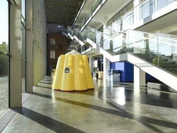 Das Puddingwunder in der Dr. Oetker Welt Gelbe Puddingform-Skulptur in modernem Glasgebäude mit reflektierendem Boden und Treppen.