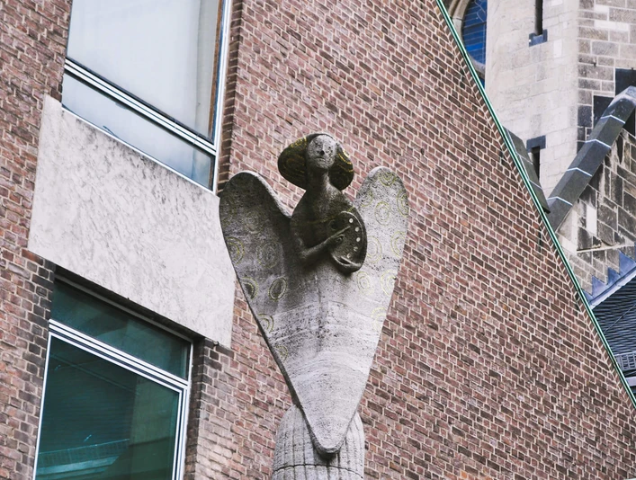 Engelsstatue Eine Engelsstatue aus Stein erhebt sich majestätisch an einem Gebäude aus roten Backsteinen.A stone statue of an angel rises majestically from a red brick building.