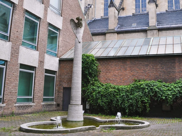 Angel statue A slender angel statue crowns a tall gray column, nestled in a courtyard surrounded by modern brick facades. It is enthroned above a small, rippling fountain that radiates tranquillity.