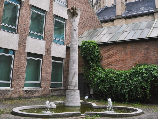 Engelsstatue Eine Engelsstatue krönt eine hohe Steinsäule, eingerahmt von Backsteinbauten und begrünten Wänden. Im Vordergrund ragt Wasser aus zwei kleinen Springbrunnen in einer modernen Wasseranlage.