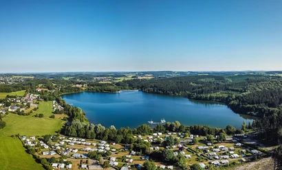 Brucher Talsperre Luftaufnahme eines großen blauen Sees umgeben von Wäldern und einer Siedlung mit Wohnmobilen.