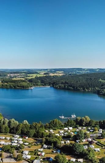 Brucher Talsperre Luftaufnahme eines großen blauen Sees umgeben von Wäldern und einer Siedlung mit Wohnmobilen.