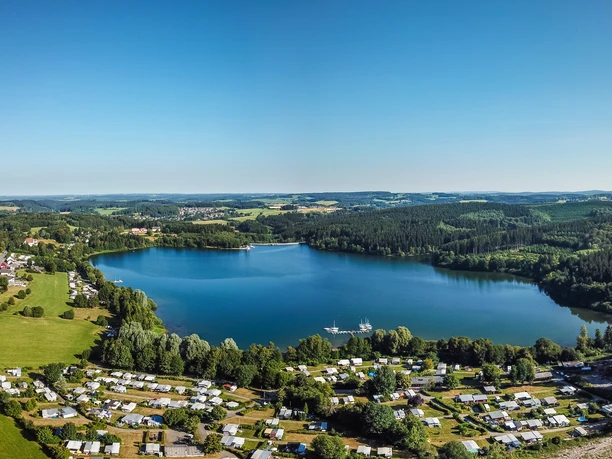 Brucher Talsperre Luftaufnahme eines großen blauen Sees umgeben von Wäldern und einer Siedlung mit Wohnmobilen.