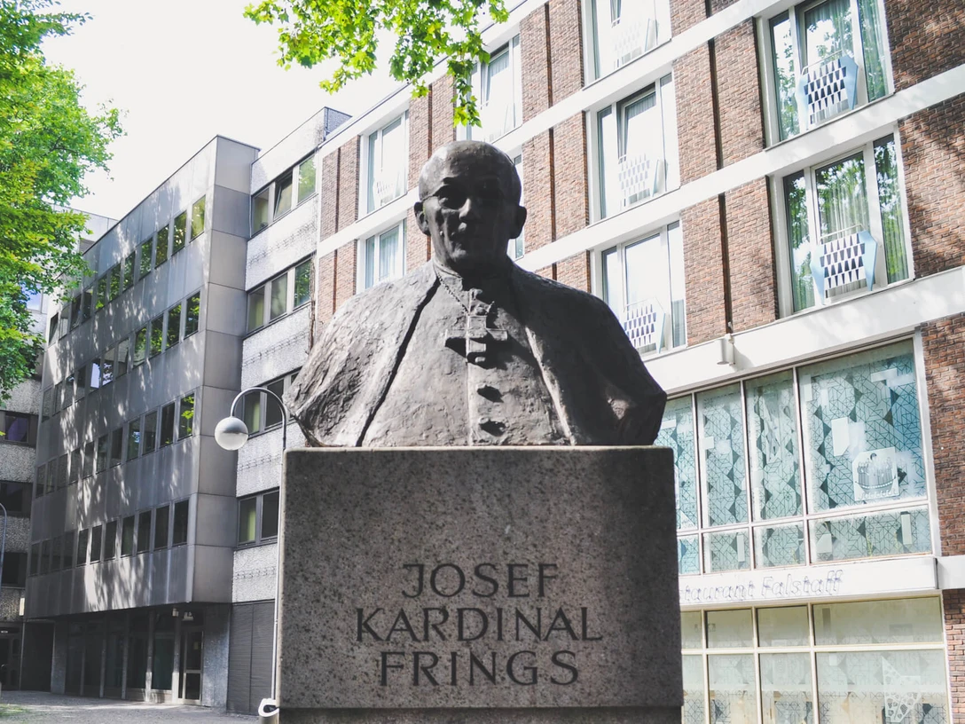 Joseph-Frings-Denkmal Das Joseph-Frings-Denkmal zeigt eine bronzene Büste des Kardinals auf einem steinernen Sockel, umgeben von modernen Gebäudeansichten.The Joseph Frings monument shows a bronze bust of the cardinal on a stone plinth, surrounded by modern views of buildings.