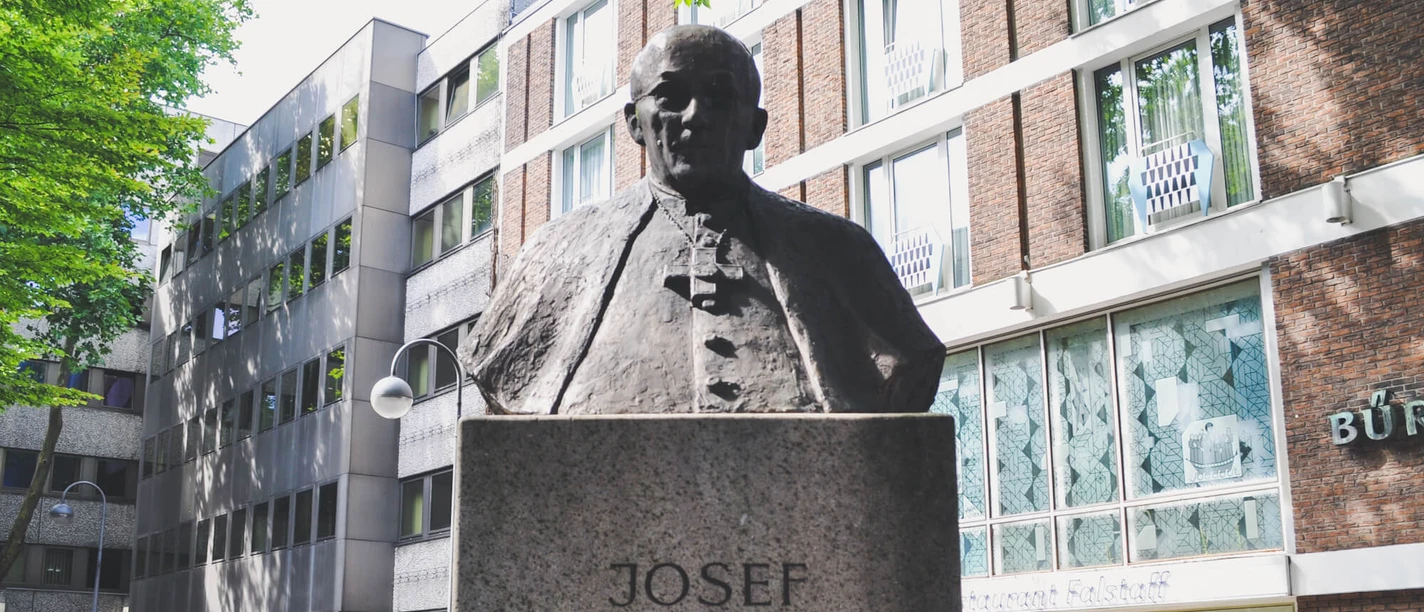 Joseph Frings Monument The Joseph Frings monument shows a bronze bust of the cardinal on a stone plinth, surrounded by modern views of buildings.