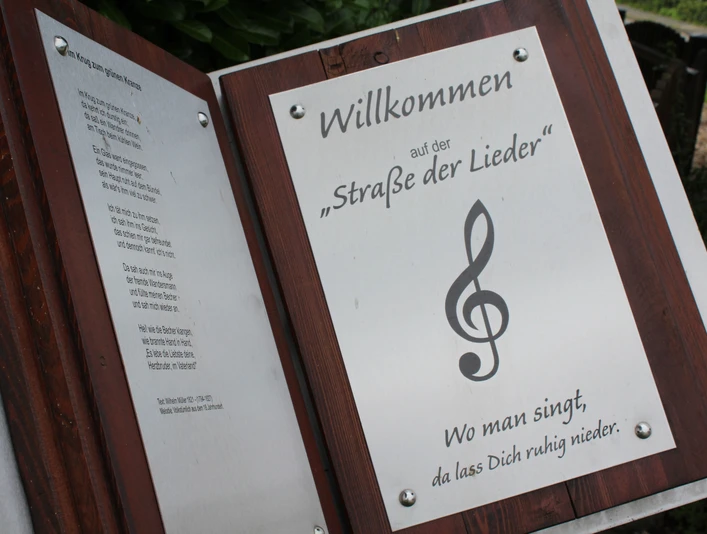 Willkommensschild der "Straße der Lieder" mit Notenschlüssel und Gedicht, inspiriert zum Mitsingen.