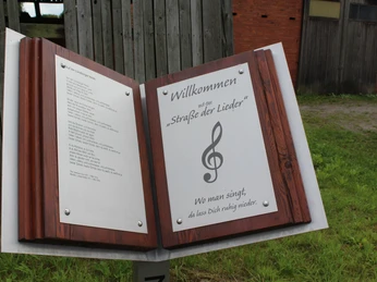 Straße der Lieder Marklohe Ein hölzernes Schild mit dem Text "Willkommen auf der Straße der Lieder" und einer Musiknote.