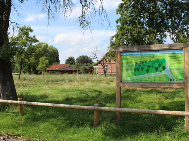 Grüne Streuobstwiese in Rodewald mit Infotafel, Bäumen und einem Bauernhaus im Hintergrund.