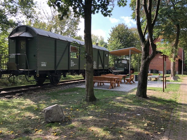A historic railroad carriage stands at a shady rest area with benches under trees.
