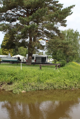 Campingplatz am Fluss mit Wohnwagen, Bäumen und Flaggen, umgeben von grüner Vegetation und Wasser.