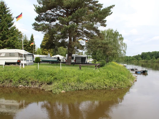Campingplatz am Fluss mit Wohnwagen, Bäumen und Flaggen, umgeben von grüner Vegetation und Wasser.