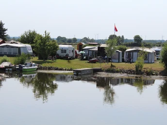 Campingplatz am ruhigen Seeufer mit Wohnwagen, Zelten und einer Fahne im Wind, umgeben von Natur.
