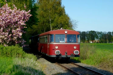 A nostalgic red train travels through a green, tree-lined landscape under a blue sky.