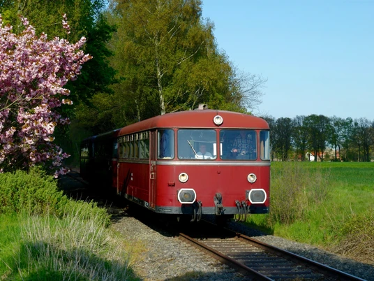 Ein nostalgischer roter Zug fährt durch eine grüne, von Bäumen gesäumte Landschaft unter blauem Himmel.A nostalgic red train travels through a green, tree-lined landscape under a blue sky.Et nostalgisk rødt tog kører gennem et grønt, træbeklædt landskab under en blå himmel.Een nostalgische rode trein rijdt door een groen, boomrijk landschap onder een blauwe hemel.