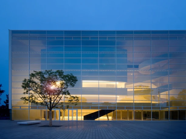 Theater Gütersloh Moderne Glasfassade eines Gebäudes bei Abenddämmerung, innen warm erleuchtet, mit Baum im Vordergrund.