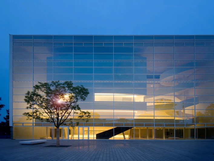 Theater Gütersloh Moderne Glasfassade eines Gebäudes bei Abenddämmerung, innen warm erleuchtet, mit Baum im Vordergrund.