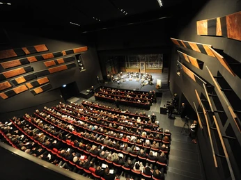 Theatersaal Moderner Theatersaal mit gefüllten Sitzreihen und einem Orchester auf der Bühne im hellen Scheinwerferlicht.