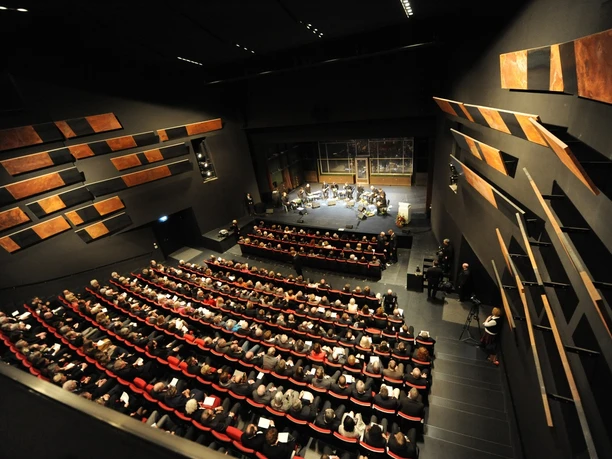 Theatersaal Moderner Theatersaal mit gefüllten Sitzreihen und einem Orchester auf der Bühne im hellen Scheinwerferlicht.