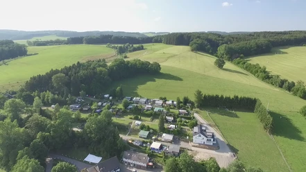 Luftbild eines idyllischen Campingplatzes umgeben von grünen Wiesen und Wäldern in hügeliger Landschaft.