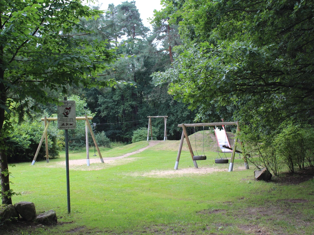 SG Steimbke - Waldspielplatz Stöckse Waldspielplatz mit Schaukel, Rutsche und Schlitten im Grünen, umgeben von hohen Bäumen und Wiesen.
