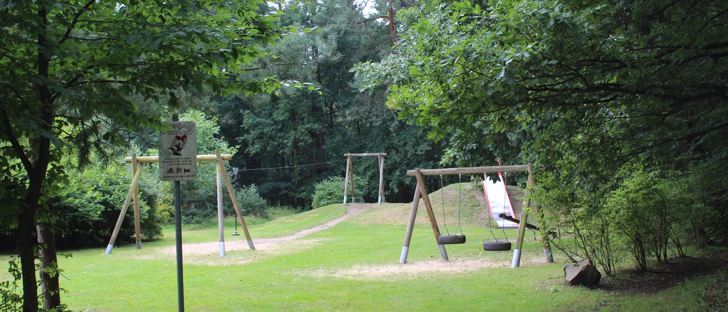 SG Steimbke - Waldspielplatz Stöckse Waldspielplatz mit Schaukel, Rutsche und Schlitten im Grünen, umgeben von hohen Bäumen und Wiesen.
