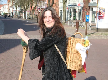 Kräuterhexe Lovis Eine Frau in schwarzem Umhang und mit Wanderstab, lächelnd auf einer gepflasterten Straße.