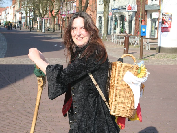 Kräuterhexe Lovis Eine Frau in schwarzem Umhang und mit Wanderstab, lächelnd auf einer gepflasterten Straße.