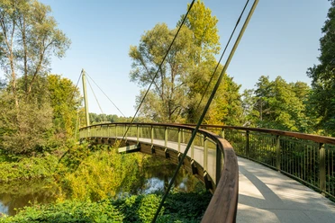 Aggerbogen Schwungvolle Holzbrücke, eingebettet in grüner Flusslandschaft unter klarem Himmel in Lohmar.