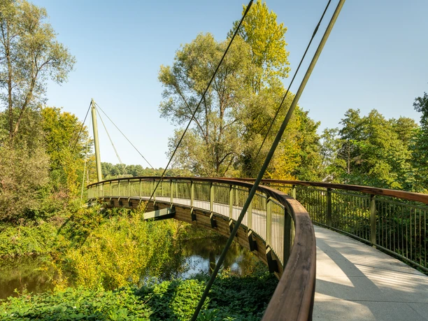 Aggerbogen Schwungvolle Holzbrücke, eingebettet in grüner Flusslandschaft unter klarem Himmel in Lohmar.