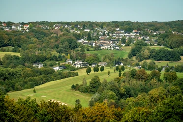 Lohmar-Wahlscheid Weitläufige Landschaft mit sanften Hügeln, grünen Wiesen und einer kleinen Siedlung am Horizont.