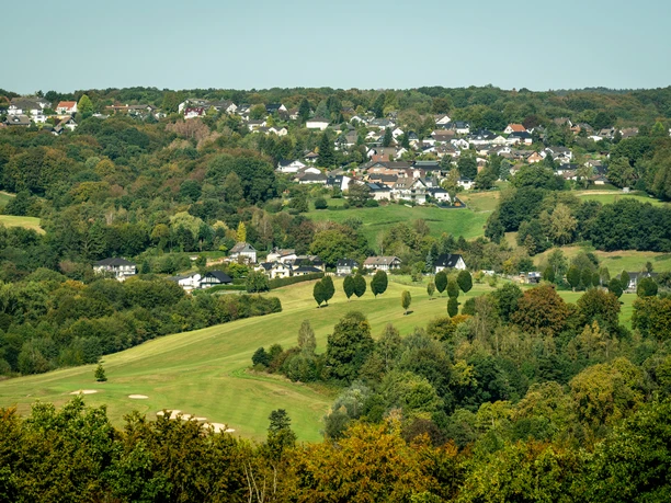 Lohmar-Wahlscheid Weitläufige Landschaft mit sanften Hügeln, grünen Wiesen und einer kleinen Siedlung am Horizont.
