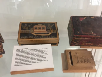 Ausstellungsstücke in der Gedenkstätte Stalag 326 VIK Holzbehälter mit Aufschriften und Symbolen, ausgestellt hinter Glas, beleuchten Kriegserfahrungen.