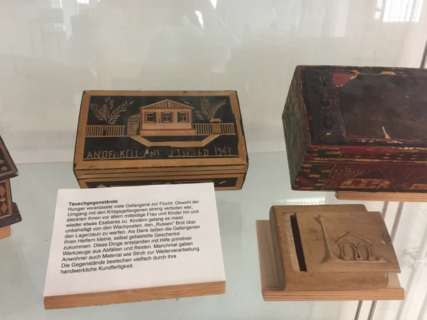 Ausstellungsstücke in der Gedenkstätte Stalag 326 VIK Holzbehälter mit Aufschriften und Symbolen, ausgestellt hinter Glas, beleuchten Kriegserfahrungen.