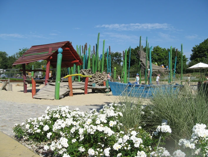 Spielplatz Schilfwald Spielplatz mit buntem Klettergerüst, Sand und Grün; Kinder spielen; weiße Blumen im Vordergrund.
