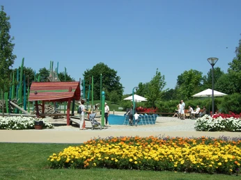 Ein blühender Park mit bunten Blumenbeeten, Spielplatz, Sitzbereichen und Spaziergängern bei Sonnenschein.