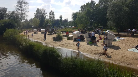 Menschen genießen einen sonnigen Tag am Sandstrand mit Strandkörben an einem kleinen See.