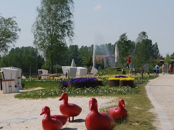 Vier rote Entenstatuen stehen auf einer Wiese, umgeben von blühenden Blumenrabatten.
