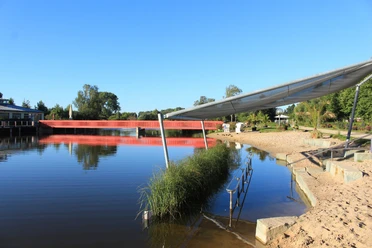 Kneippbecken Auf dem Bild ist ein See mit roter Brücke, Sandstrand und grüner Umgebung unter klarem Himmel.